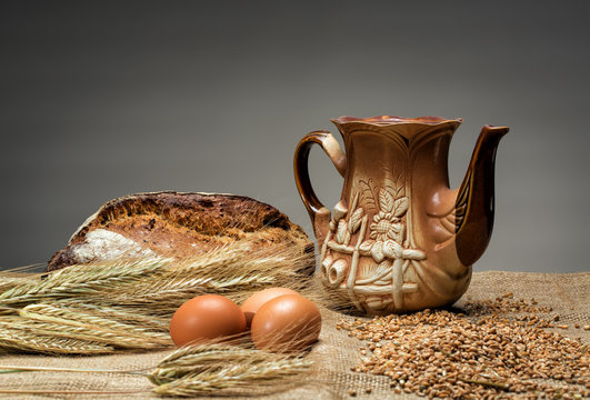 Jug, bread, eggs and spikelets on burlap on a gray background