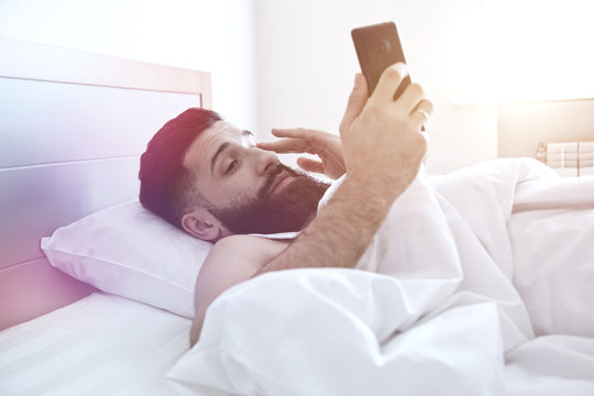 Sleepy man lying in morning bed with phone using app or reading news feed