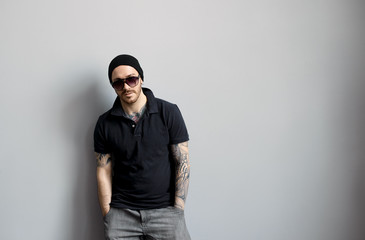 Style and fashion concept. Studio shot of trendy handsome bearded young man wearing stylish sunglasses and hat having confident look posing at grey wall, keeping hands in pockets of his jeans