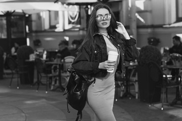 Obraz na płótnie Canvas Young beautiful stylish woman wearing green dress, black jeans jacket and sunglasses walking at the street in city. Pretty brunette girl with long hair holding backpack and coffee