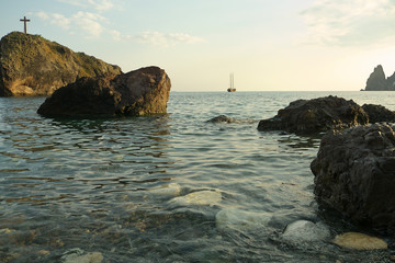 Landscape view of rock with cross and stones on the empty beach, yacht in the sea at background