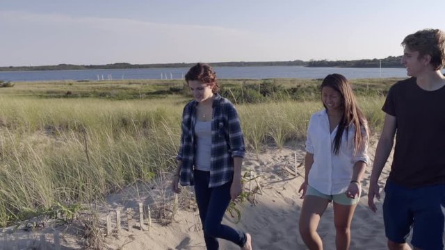 Multiethnic Teens Go For A Walk On Sandy Beach Path, Salt Water Pond In Background