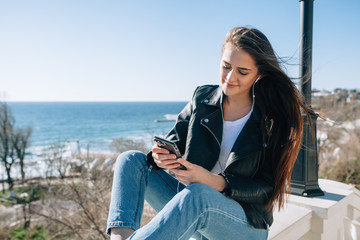 The beautiful girl with dark long hair which are developed by wind sits by the sea. She is keen on something in phone, beams of the sun force to smile the girl.