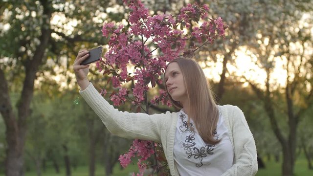 A girl makes selfie in the garden. An attractive red-haired woman smiles making selfi using a mobile phone in a cherry orchard. The concept of using gadgets for a healthy lifestyle. Young sporty happy