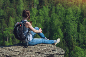 Girl with backpack sitting on a rock on top of a mountain on the background of green forest