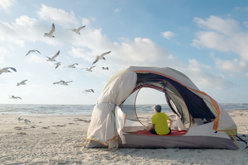 A teenager in a tent on the shores of the Gulf of Mexico looks at flying seagulls. Padre Island National Seashore, Texas, United States