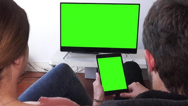 Young Couple Watching Green Screen Devices Relaxed At Home. Couple watching television and tablet green screen. Close up shot behind models shoulders