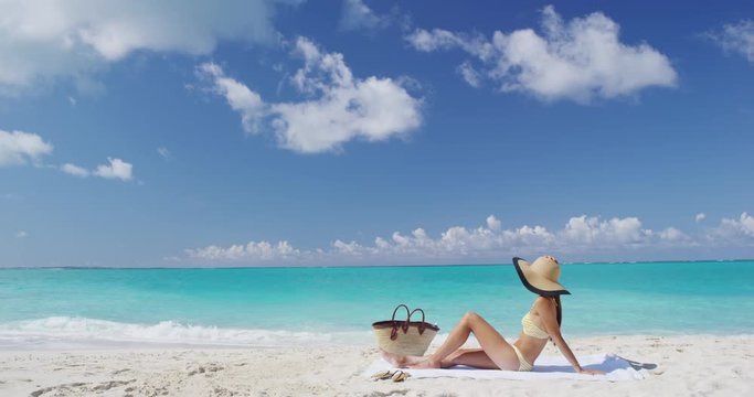 Sexy bikini woman sunbathing relaxing on beach. Suntan concept. Young adult lying down with straw hat sun tanning under the tropical sun on Caribbean vacation.