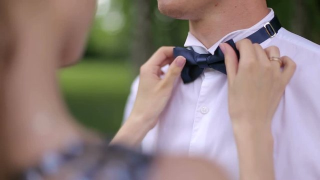 Girl corrects her husband a bow-tie round his neck in the outdoors, close-up. Young woman helping man with bow-tie.