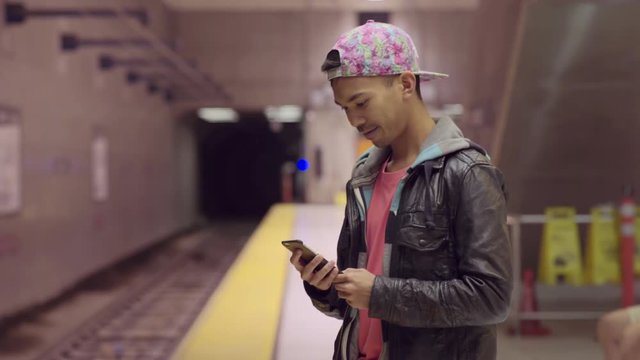 Asian Man Uses Smart Phone While Waiting For Subway Train, Motions For His Boyfriend To Join Him
