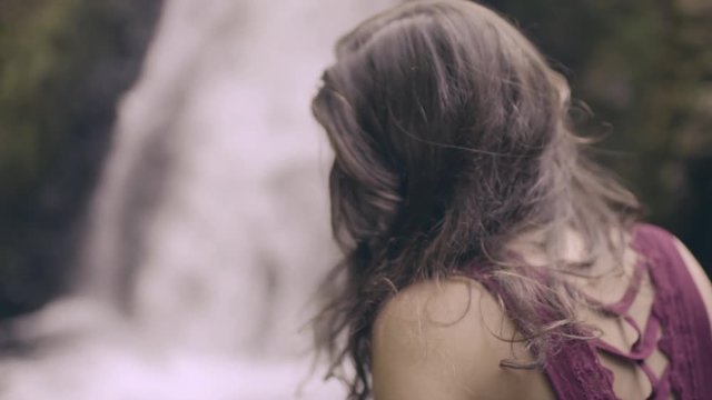 Young Woman Sits On A Cliff Across From Waterfall, Camera Gets Closeup Of Her Feet Dangling Over The Edge For Perspective (Slow Motion)
