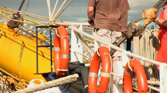 Visitor at bow of tall ship looks out over bowsprit of Dewaruci holding onto railing with three bright orange life-rings.   