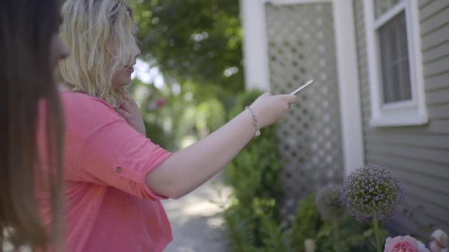 Girl Takes A Photo Of A Large Purple Flower (Allium) Growing In A Garden, Her Friend Touches The Flower (Slow Motion)
