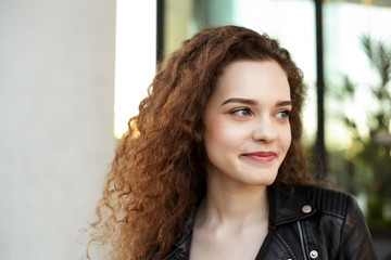 Oudoor urban portrait of cute beautiful young European female with curly hair and charming smile breathing fresh air during evening walk on city streets, standing against modern building background