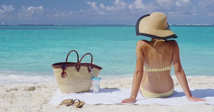 Vacation travel concept on Beach - Woman in bikini sun tanning relaxing on beach. Unrecognizable girl from the back sitting down with straw hat sunbathing under the tropical sun on Caribbean vacation.