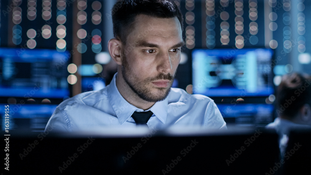 Poster Close-up of a Professional Technical Controller Sitting at His Desk with Multiple Displays Before Him. In the Background His Colleagues Working in System Control Center.