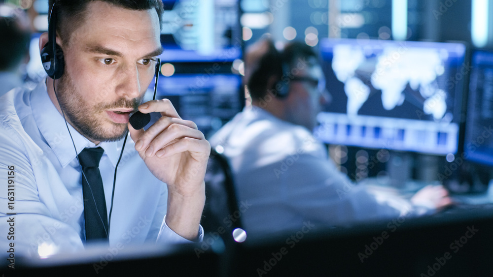 Poster in monitoring room technical support specialist speaks into headset. his colleagues are working in t