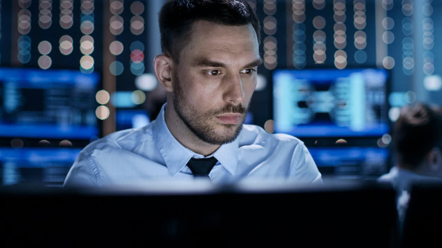 Close-up of a Professional Technical Controller Sitting at His Desk with Multiple Displays Before Him. In the Background His Colleagues Working in System Control Center. - Powered by Adobe