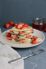 Delicious pancakes on grey table with fruits. Plate full of pancakes with strawberries, bananas and chocolate cream. Pouring over by Syrup. Berry jam in glass jar
