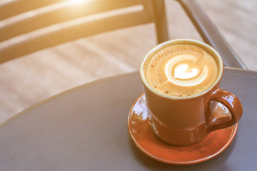 Hot latte coffee cup on black metal table in coffee shop with wooden floor and sunlight background.