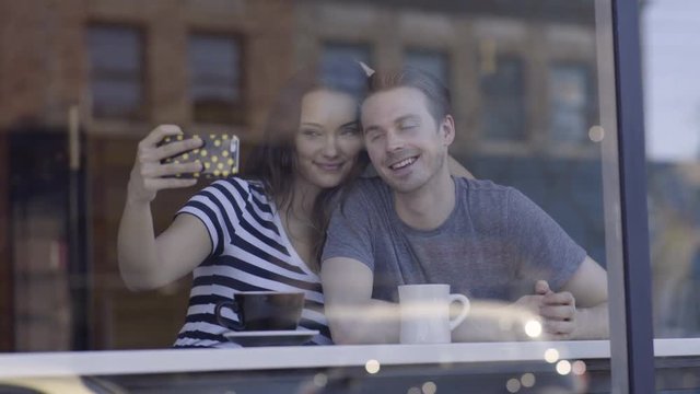 Fun Couple Take Selfies Together In Coffee Shop Window, Boyfriend Makes A Silly Face To Make His Girlfriend Laugh