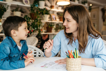 Mom and son painted with colored pencils in a cozy room, look at each other and smile. Shelves with flower pots on the backgrounds