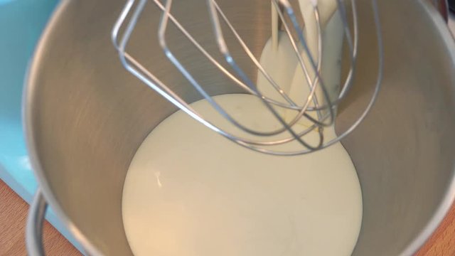 Milk is poured into a steel bowl with a whisk of an electric beater above - closeup from above