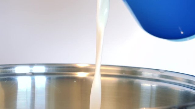 A woman pours milk from a blue plastic measuring jug into a steel bowl - closeup
