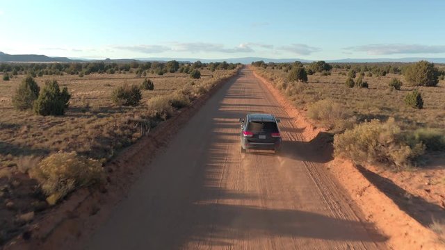 AERIAL: Black SUV car driving along the empty dusty road trough dry meadow desert valley in Utah on sunny morning. Flying above jeep traveling on straight dirt route into Capitol Reef at golden sunset