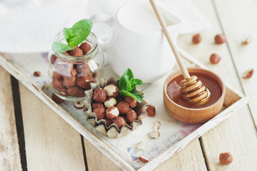 Honey in the wooden bowl, mint leaves, hazelnuts and jar with milk on the wooden tray