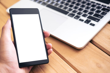Mockup image of hand holding black mobile phone with blank white screen and silver color laptop on vintage wooden table background