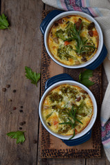 Baked casserole from eggs, bacon, dried tomatoes, broccoli and cheese in blue ramekin on brown wooden background. Mushroom gratin. Breakfast or lanch
