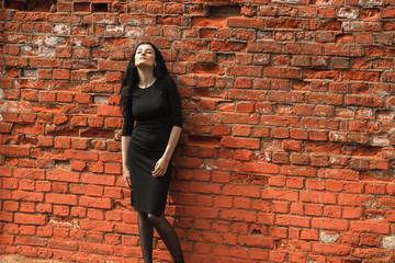 Fototapeta na wymiar Portrait of young beautiful white girl with aristocratic features with long black hair and in black clothes posing near vintage red brick wall on a summer street.
