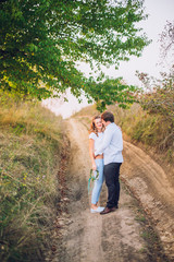 Fototapeta premium A loving couple are standing in the field road with trees at sunset, holding hands and kissing each other. Bright sunlight shines. Around the greens. Walk on the nature. Country style.