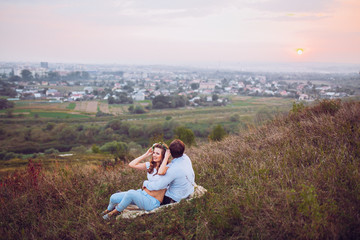 Naklejka na ściany i meble couple, love, nature and people concept - happy loving couple sitting on a plaid in a field on a background of magical sunset