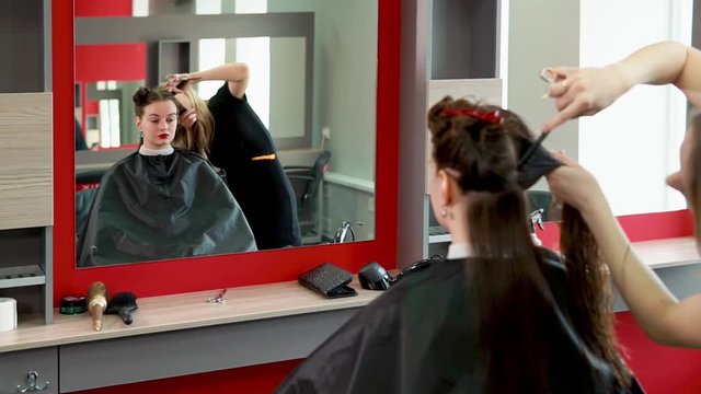 A blonde hair stylist strips wet hair into sections in order to make a haircut, a visitor to a salon with chestnut hair awaits a change of image