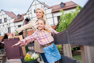 happy mother and daughter with shopping bags standing with arms outstretched near fence on street