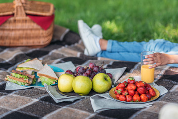 Close-up view of fresh fruits and sandwiches on plaid and person drinking juice behind