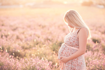 Fototapeta na wymiar Pregnant woman 24-29 year old holding belly wearing dress with floral pattern in lavender field. Posing outdoors. Motherhood. Maternity.