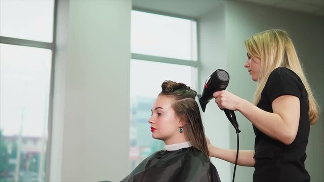 The blonde hairdresser dries long hair with the help of a warm air dryer and comb, a woman who came to the beauty salon expects a change of image and good service