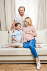 Fototapeta na wymiar Happy pregnant woman with daughter sitting on sofa and smiling man standing behind