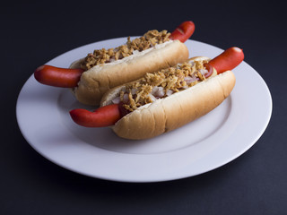 Two Danish hot dogs with red sausage, ketchup, mustard sauce and raw and fried onions on white plate, isolated on black background