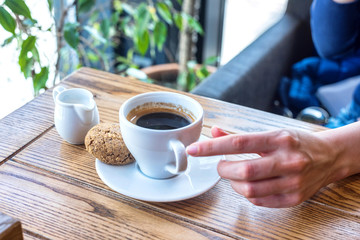 Coffee cup with cookie on wooden table in restaurant with view on window and plant. Woman touching cup with hands to drink.