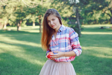 Portrait of beautiful young Caucasian woman with long red hair in plaid shirt and pink tutu tulle skirt,  in park meadow at summer sunset, looking in camera, holding apple