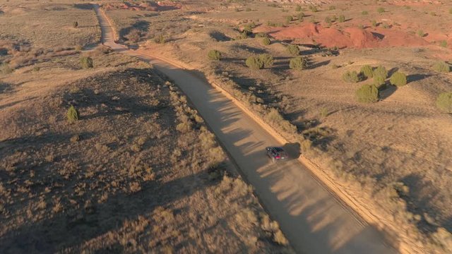 AERIAL: Black SUV car driving along the empty dusty road trough dry meadow desert valley in Utah on sunny morning. Flying above jeep traveling on straight dirt route into Capitol Reef at golden sunset