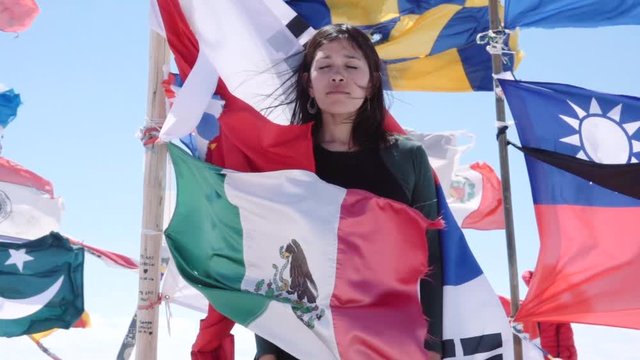 Hispanic woman stands between the countries flags waving with the wind on Uyuni salt flats. Slow motion