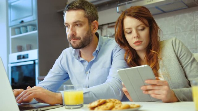 Caucasian young couple siting in kitchen. Young pretty woman using tablet while her husband using laptop. Indoor.