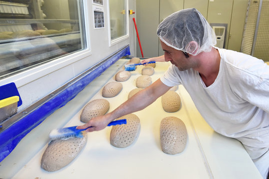 Mann mit Haarnetz und Arbeitskleidung in einer Großbäckerei bestreicht Brotlaibe am Fliessband 