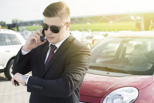 Modern businessman standing in a parking lot. Making a phone call while looking at his watch. Lens Flare in the background.
