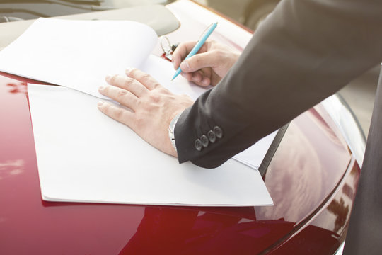 Closeup of a man's hand filing a blank paper form on a clipboard. Modern businessman standing in a parking lot leaning on a hood of the car. Lens Flare in the background.